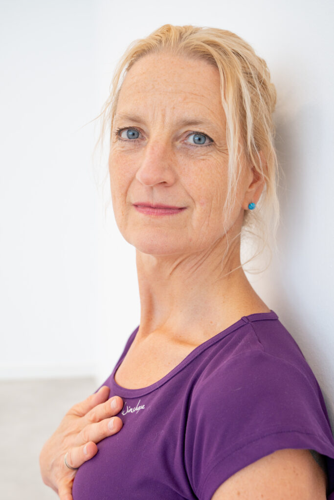 Miriam Wessels im lila Shirt, die Hand auf der Brust, seitlich an eine helle Wand gelehnt